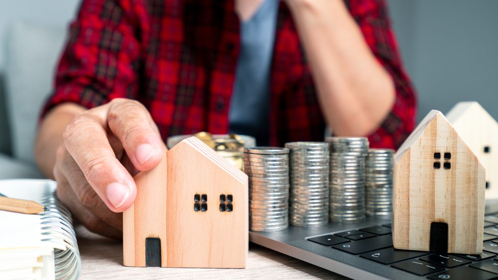 muž v košili drží na stole dřevěný model domu s hromadou mincí, koncept spoření na nový dům